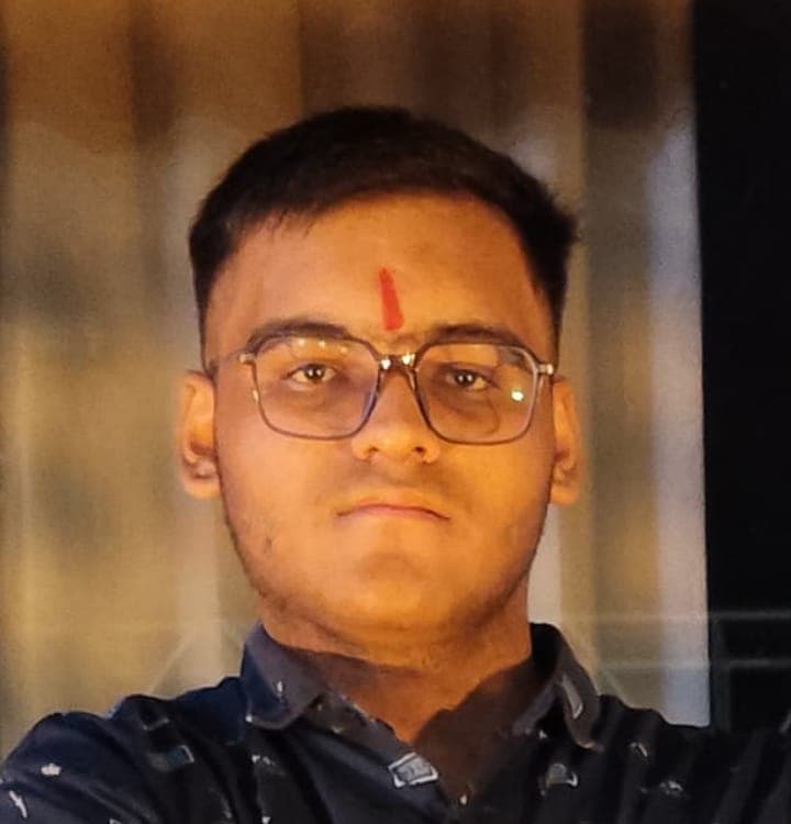 Young man with glasses and red tilak on forehead wearing a dark patterned shirt.